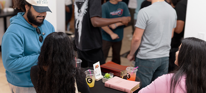 Several people gathered around a table with two women seated, a man in a blue hoodie and cap standing nearby, and others standing and talking in the background; the table has drinks, a small green plant, and a pink book titled 'Holy Bible'.