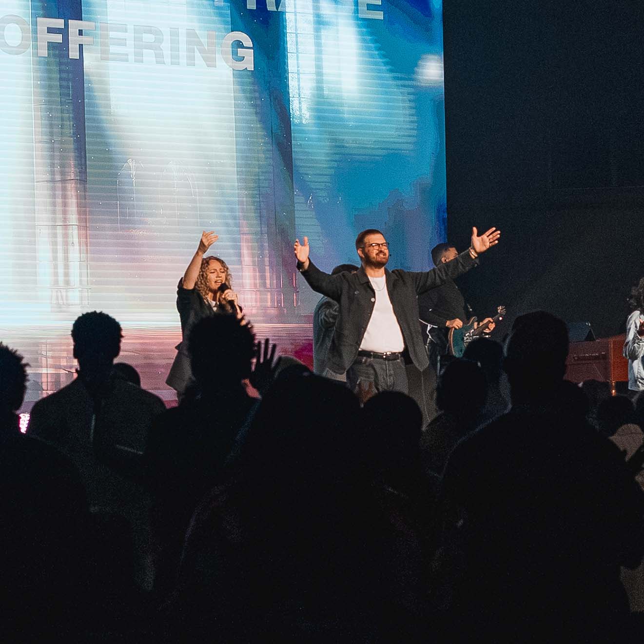 Worship band performing on stage with a female singer holding a microphone and a male preacher with glasses raising his arms, while a guitarist plays in the background and an audience watches with raised hands.