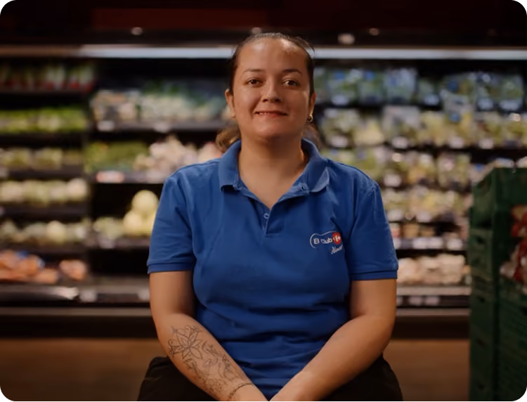 Smiling woman in a blue polo shirt with a tattoo on her forearm sitting in a grocery store produce aisle.