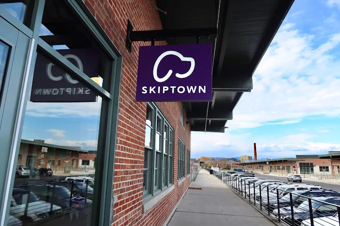 Purple sign with white cloud outline and text 'SKIPTOWN' mounted on a brick building along a sidewalk with parked cars on the right.