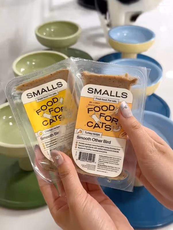 Two hands holding Smalls brand cat food packets labeled 'Smooth Bird' and 'Smooth Other Bird' with stacked green and blue cat bowls in the background.