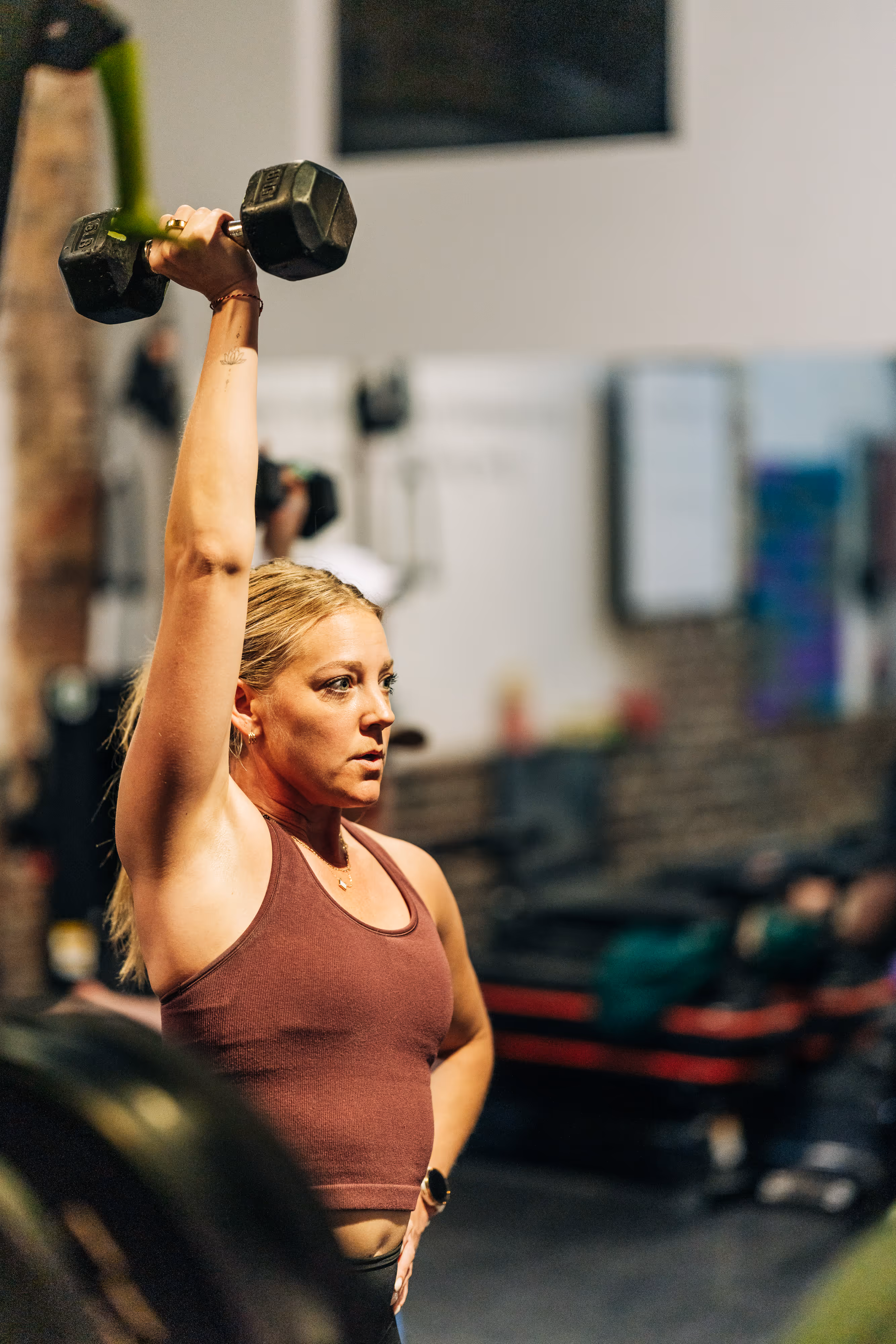 Woman in a gym lifting a dumbbell overhead with a focused expression.