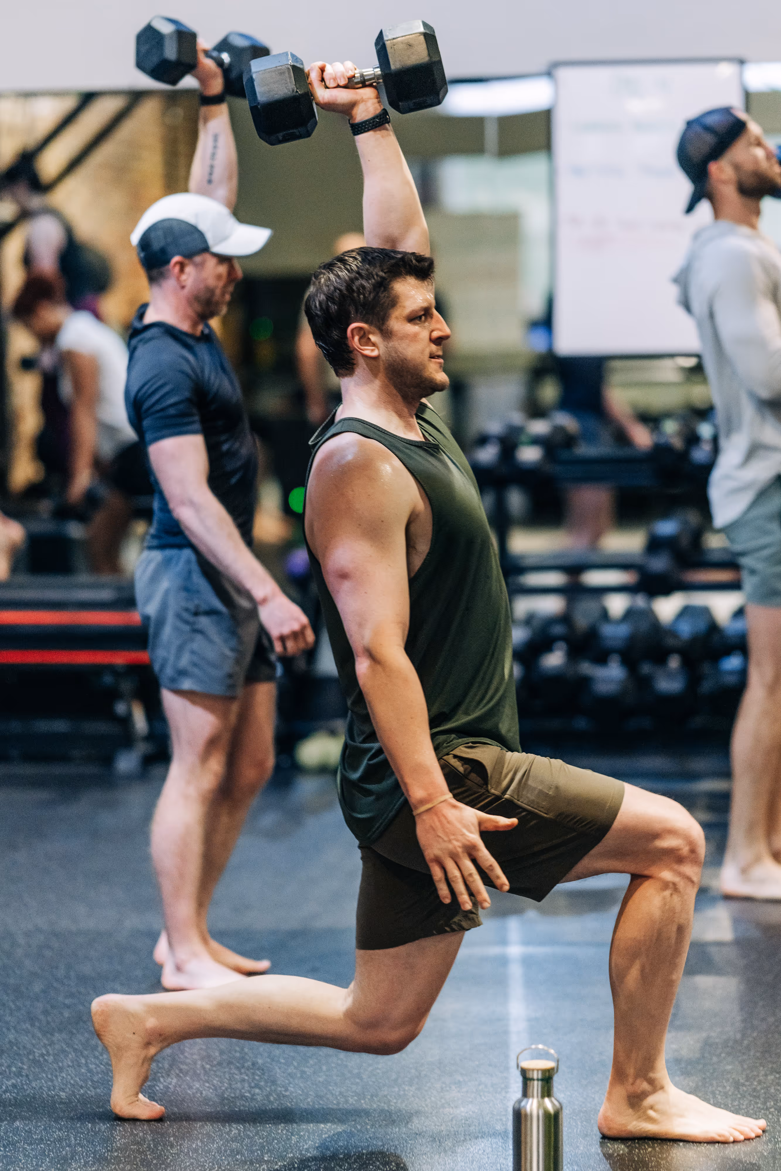 Man in sleeveless shirt and shorts performing overhead dumbbell lunge in a gym with others exercising in the background.