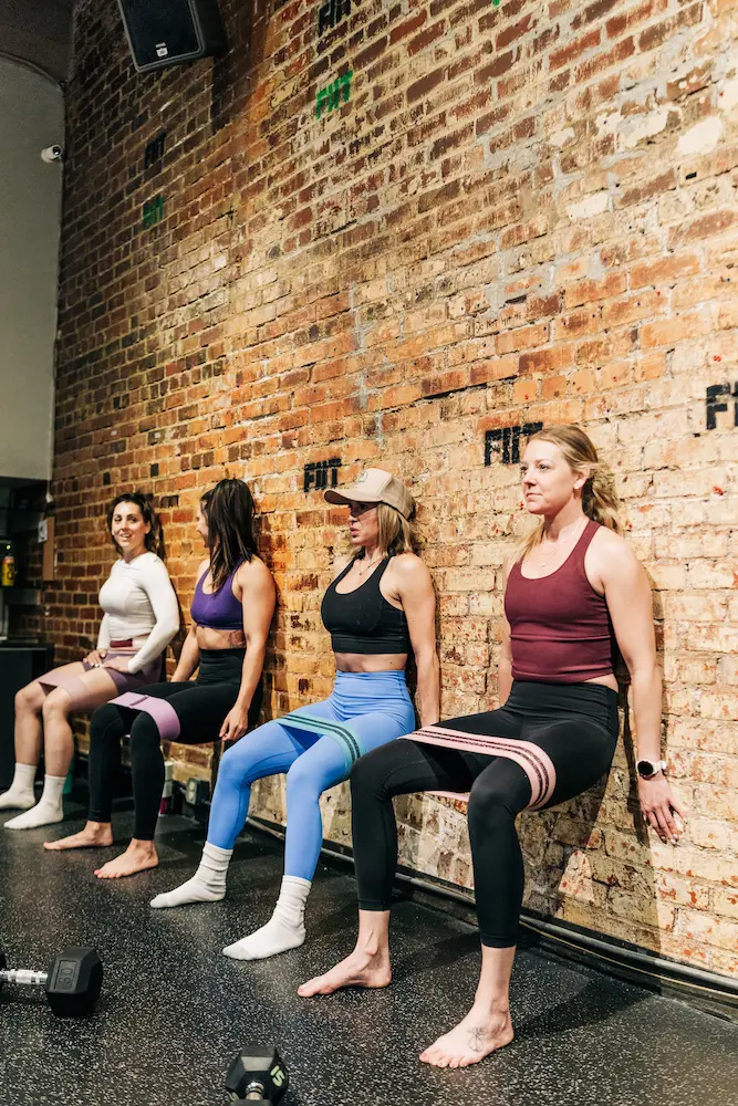 Four women performing wall sits in a gym using resistance bands around their thighs.