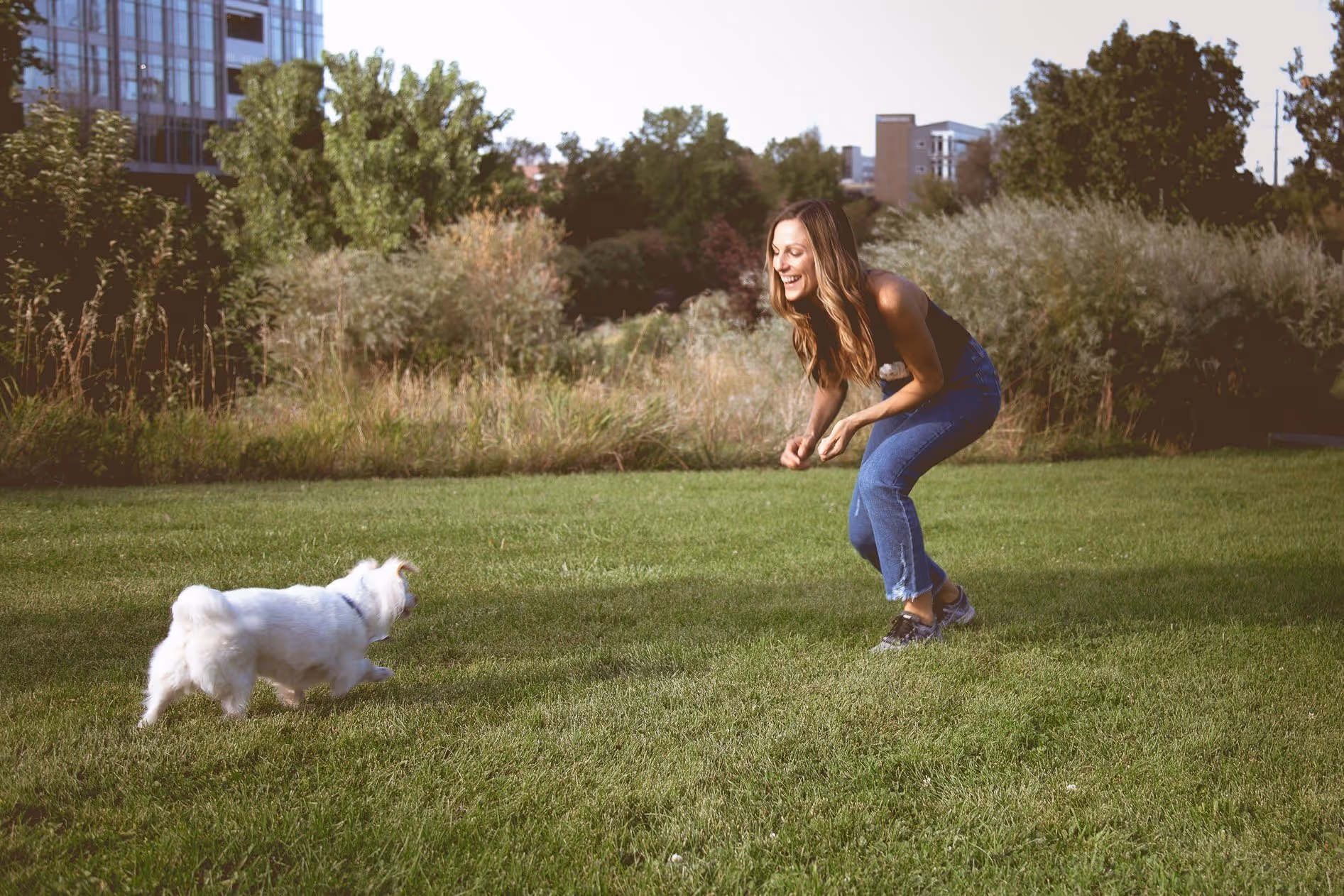 Woman crouching and smiling while playing with a small white dog on green grass in a park.