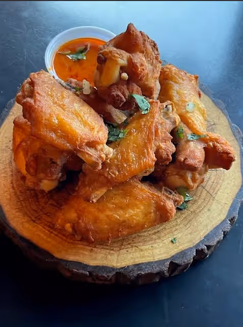Crispy fried chicken wings garnished with herbs, served on a wooden plate with a small cup of dipping sauce.