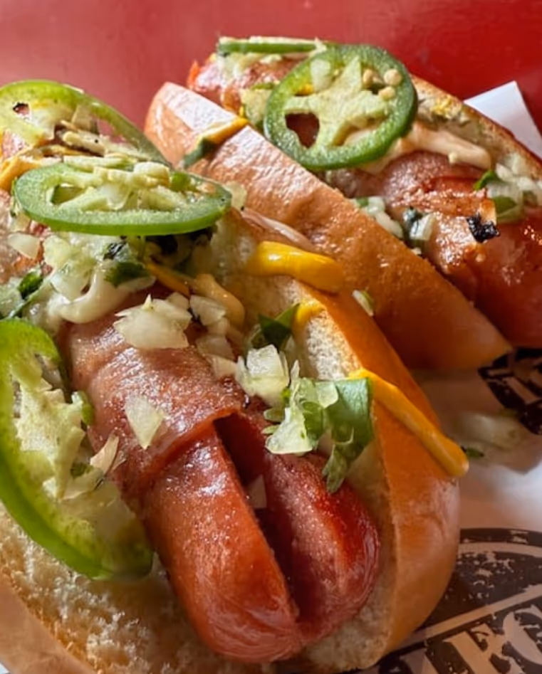Two hot dogs in buns topped with jalapeño slices, chopped onions, shredded cheese, and chopped green herbs on a red background.