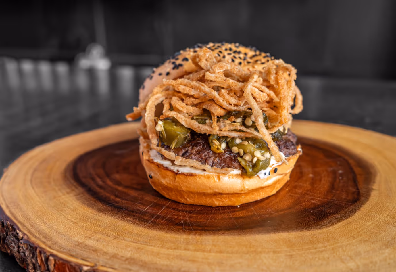 Burger with fried onion strings, jalapeños, and beef patty on a sesame seed bun, placed on a wooden serving board.