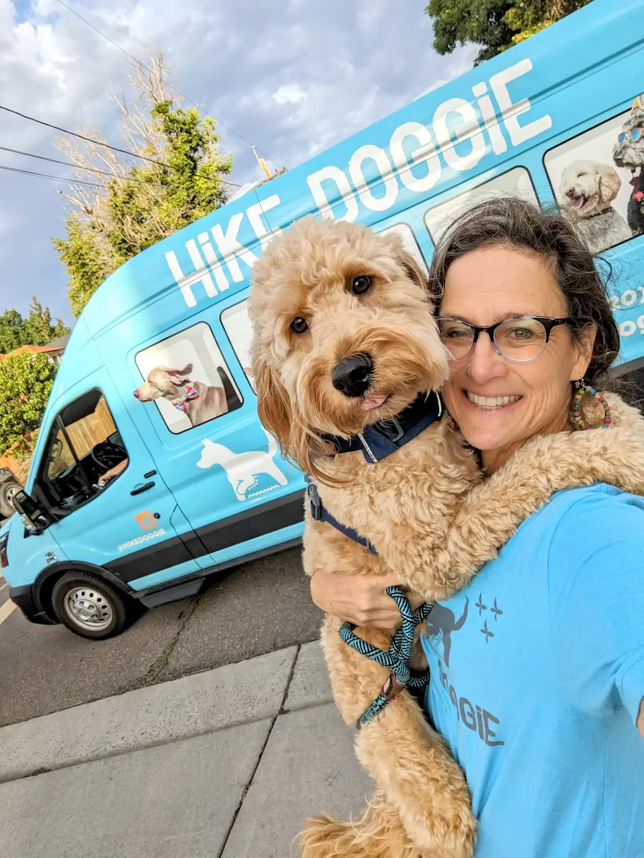 Woman holding dog in front of blue Hike Doggie van for urgent vet care in Denver.