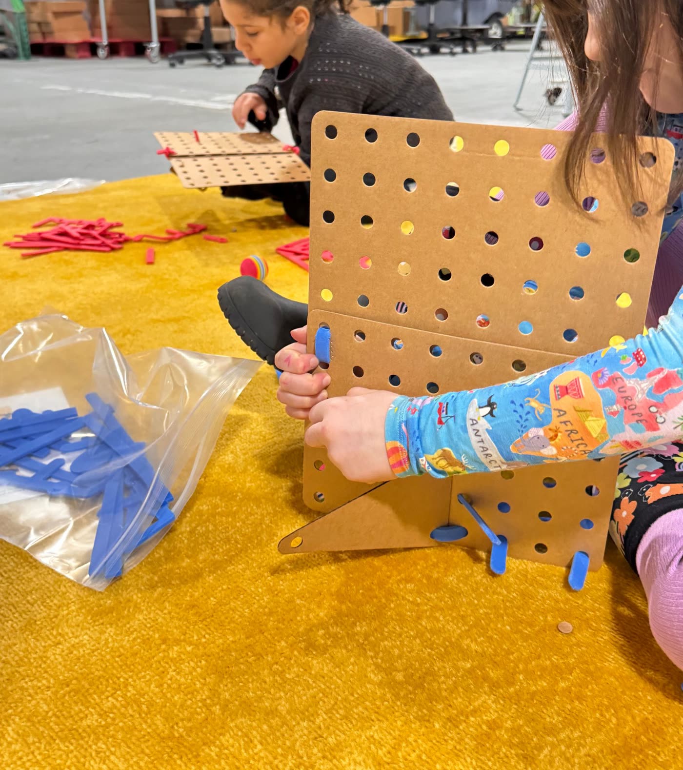 Two learners building Mazes Sparks on a yellow rug, one fitting foam pieces into a cardboard pegboard
