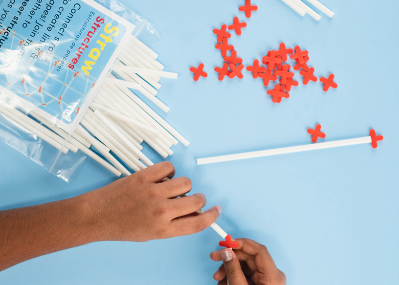 Overhead close-up of two hands assembling a Straw Structures kit, with white straws and red plastic connectors visible on a blue background.