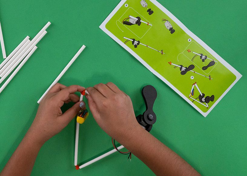 Overhead close-up of two hands assembling a Flying Machines wind-up motor onto a straw frame, with the printed instruction card visible on a green background.