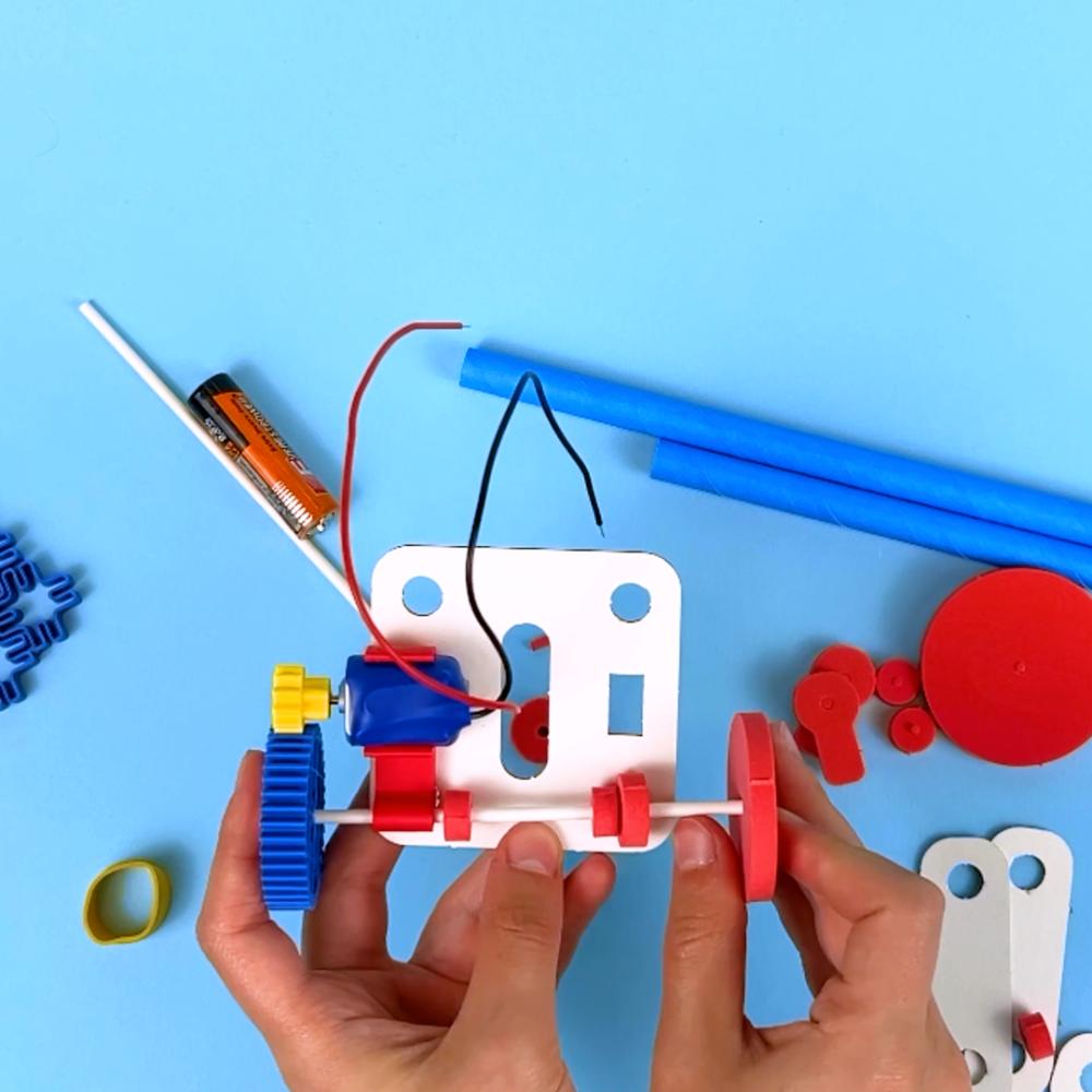A pair of hands assembling an Electric Vehicles chassis, showing the motor, battery, gears, and axle on a blue background.