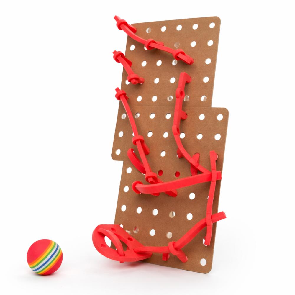 An assembled cardboard Mazes board standing upright with red foam ramps zig-zagging down through die-cut holes to a hoop at the bottom, and a rainbow-striped ball resting beside it