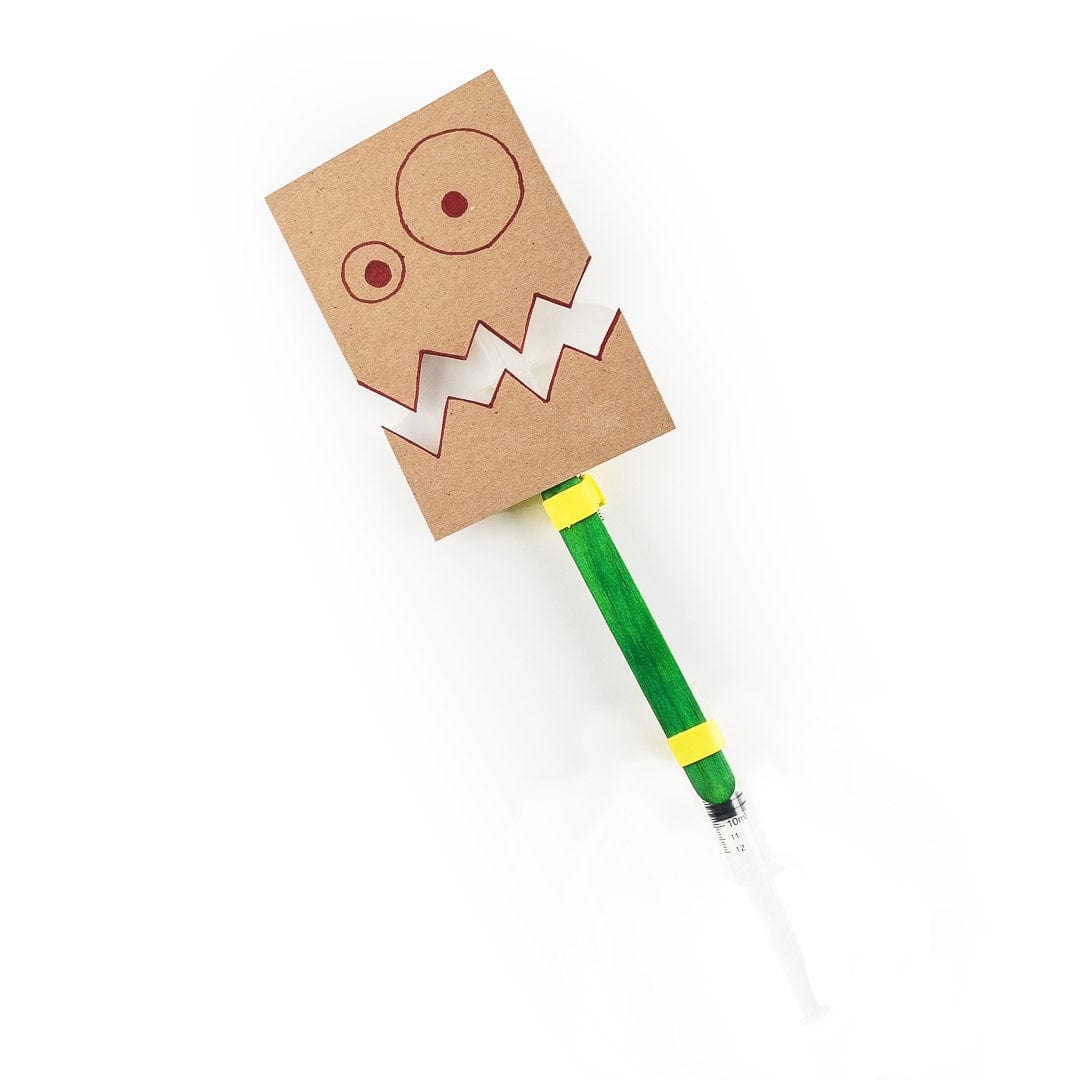 A rectangular cardboard monster face with two hand-drawn eyes and a jagged mouth, mounted on a green-decorated craft stick above a syringe-style piston and cylinder