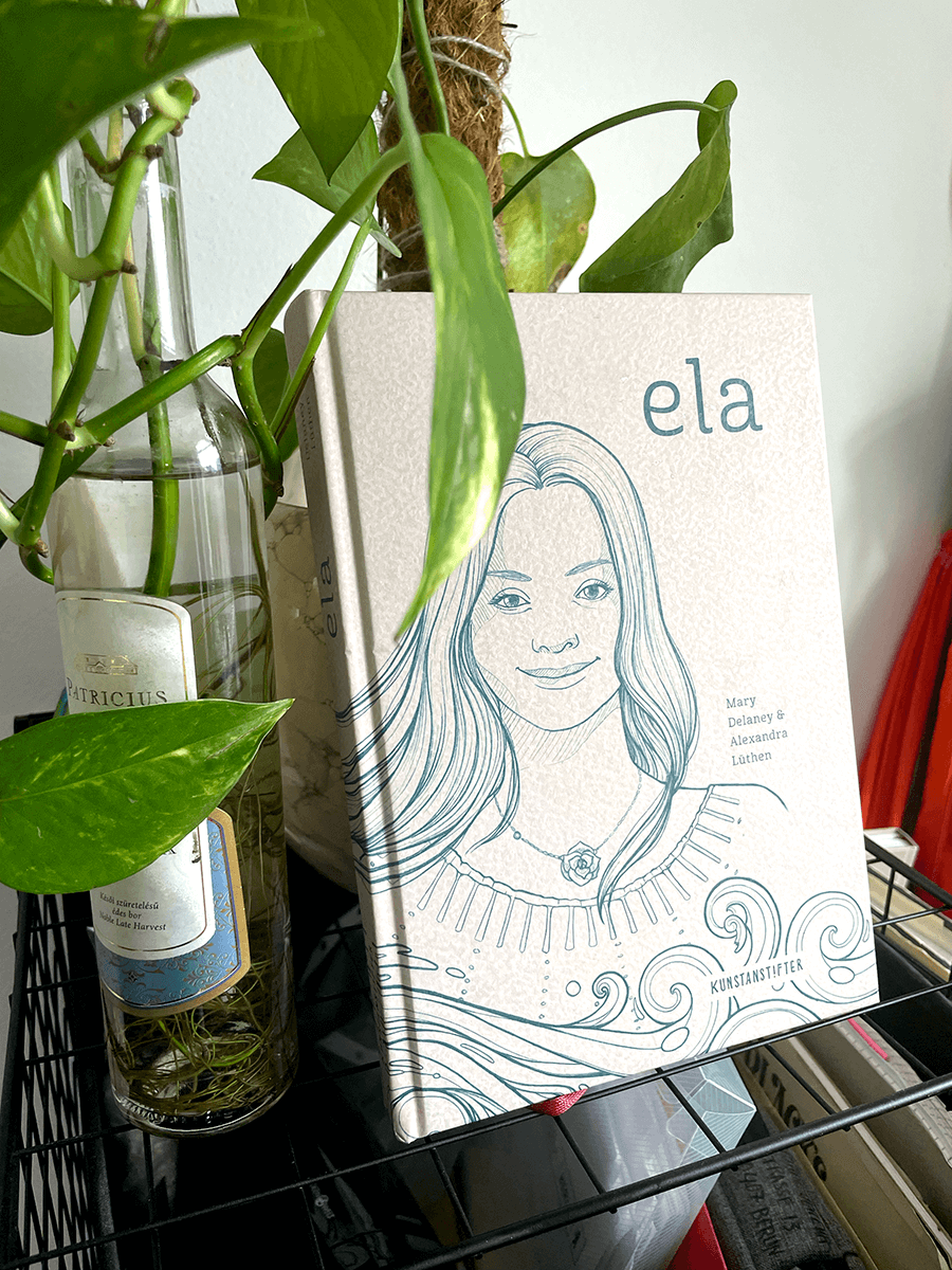 Ein Foto des Buches Ela neben den Blättern einer Pflanze. Das Buchcover zeigt den Titel und eine Abbildung des Gesichts einer Frau mit langen, wirbelnden Haaren und kleinen Wirbeln, die das Meer darstellen. 