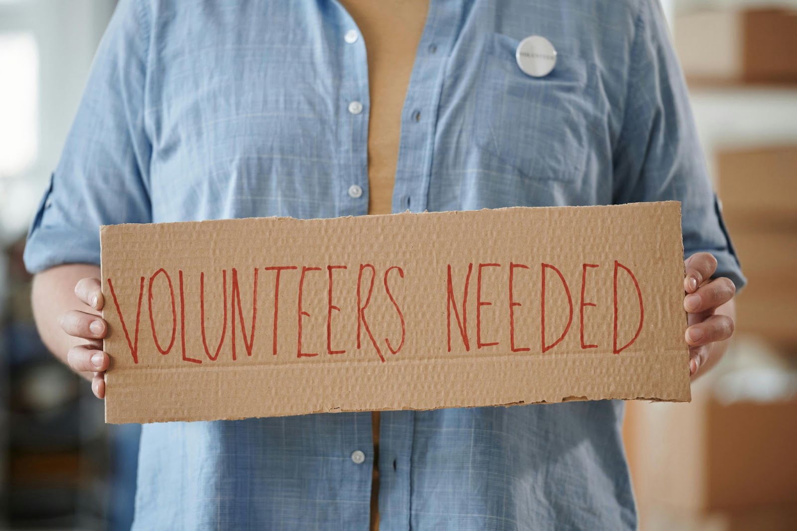 Person holding a cardboard sign that reads “Volunteers Needed” to encourage community involvement.‍