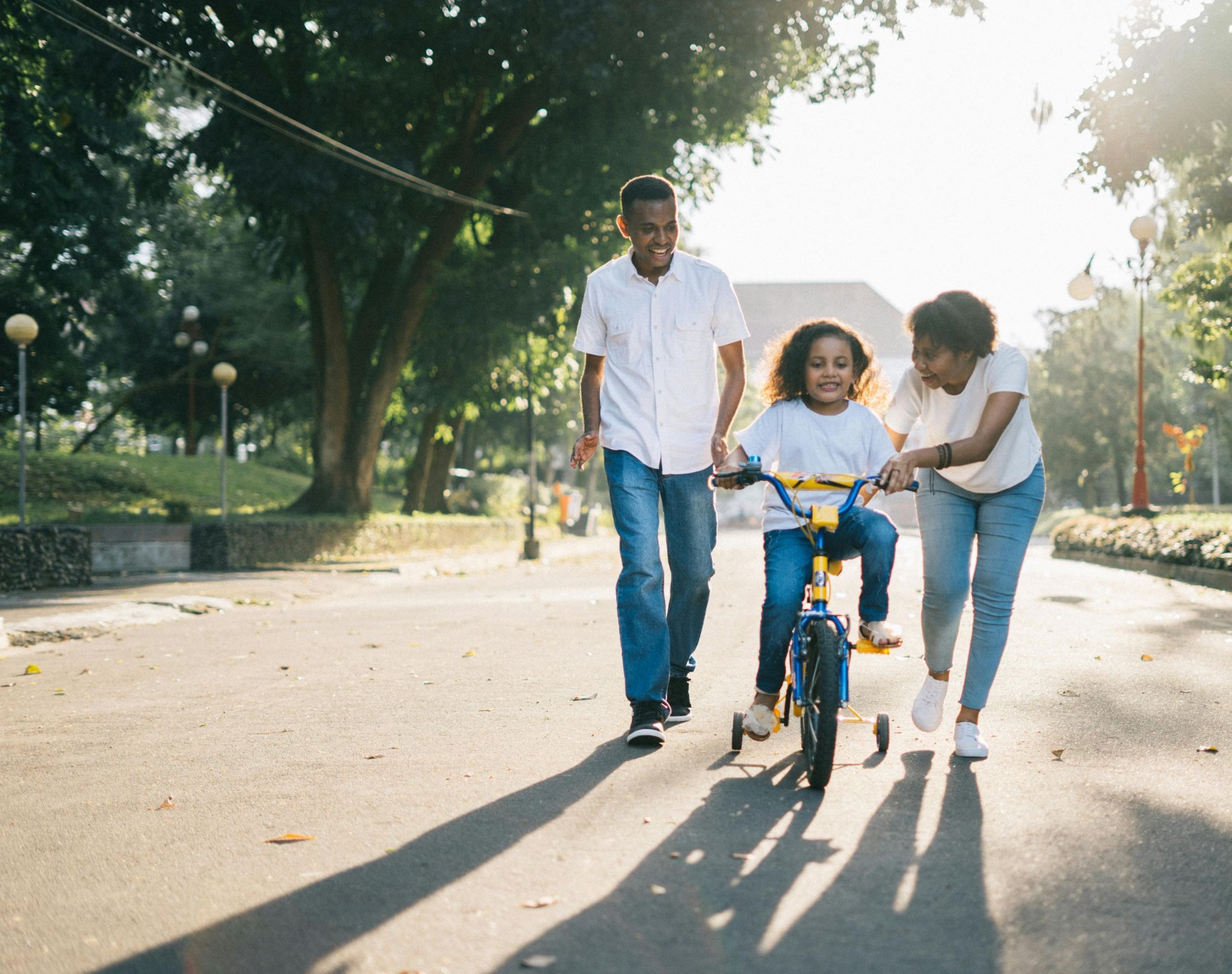 Supportive parents smile as they help their young daughter learn to ride a bicycle in a sunny park.