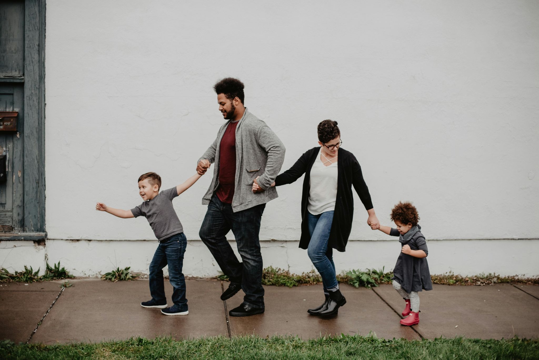 A happy, diverse adoptive family with two parents and two young children walk together holding hands along a sidewalk.