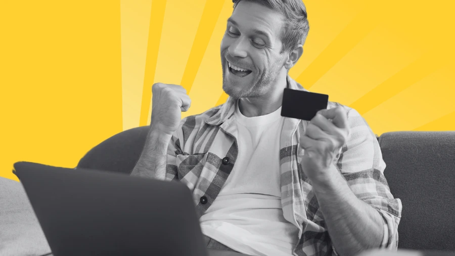 A man sits on a couch, smiling and raising his fist in excitement while holding a credit card in one hand and looking at a laptop. The background is bright yellow with sunburst patterns.
