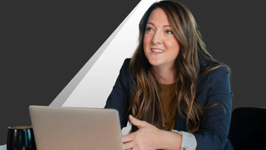 A woman with long brown hair, wearing a blazer, sits at a table with a laptop and a mug, smiling and gesturing with her hand against a dark, geometric background.