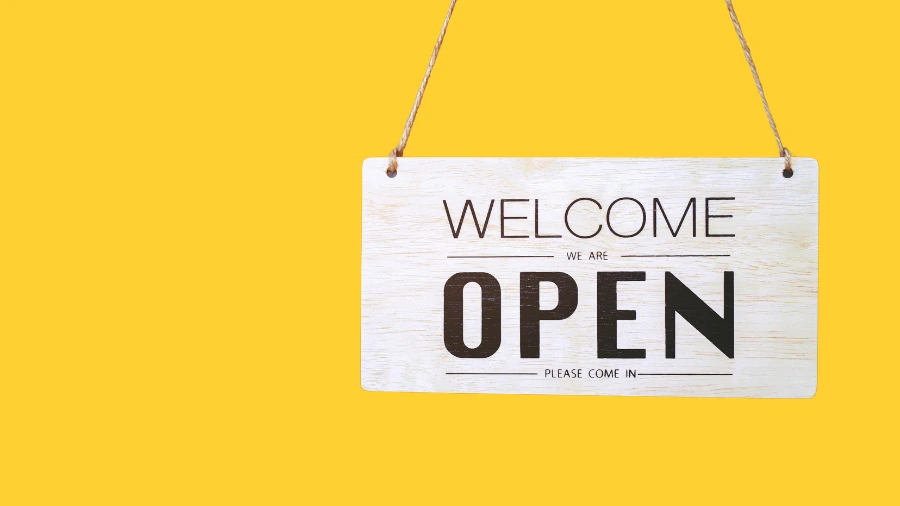 A wooden sign hanging on a yellow background reads Welcome, We are OPEN, Please come in in bold black letters.