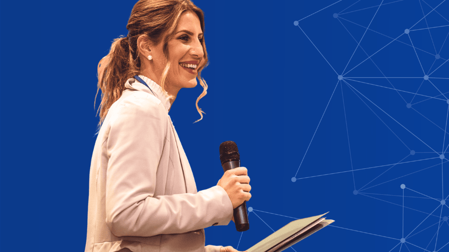 A woman with wavy hair in a ponytail, wearing a light blazer, holds a microphone and papers while smiling, standing in front of a blue background with a network graphic pattern.