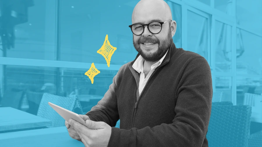Smiling man with glasses holds a tablet while seated outdoors, with a blue graphic background and yellow spark icons.