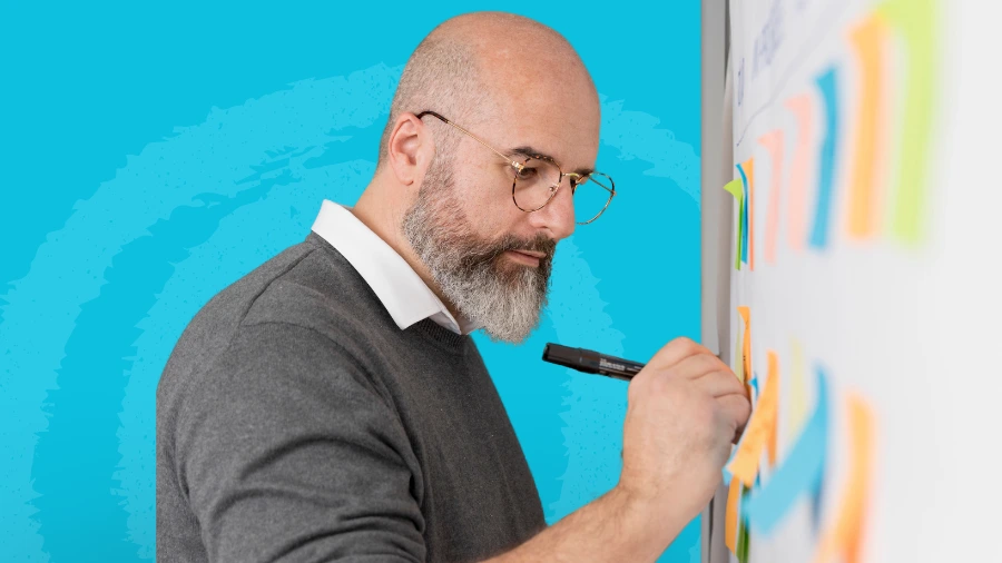 Bald man with glasses writing on a whiteboard covered with colorful sticky notes against a blue background.