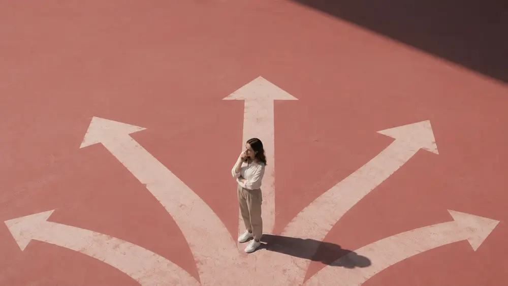 Aerial-view conceptual thumbnail of a woman standing among multiple directional arrows on a muted red surface, symbolizing hesitation and decision-making before booking