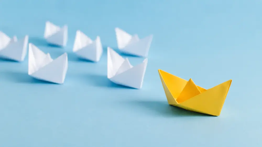 Minimalist scene featuring a yellow paper boat standing apart from several white paper boats on a soft blue background, symbolizing trust, distinction, and perceived value among similar choices.