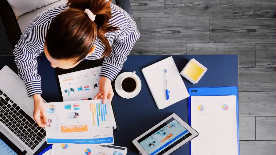 Business professional reviewing charts and reports at a desk with a laptop, tablet, and coffee.