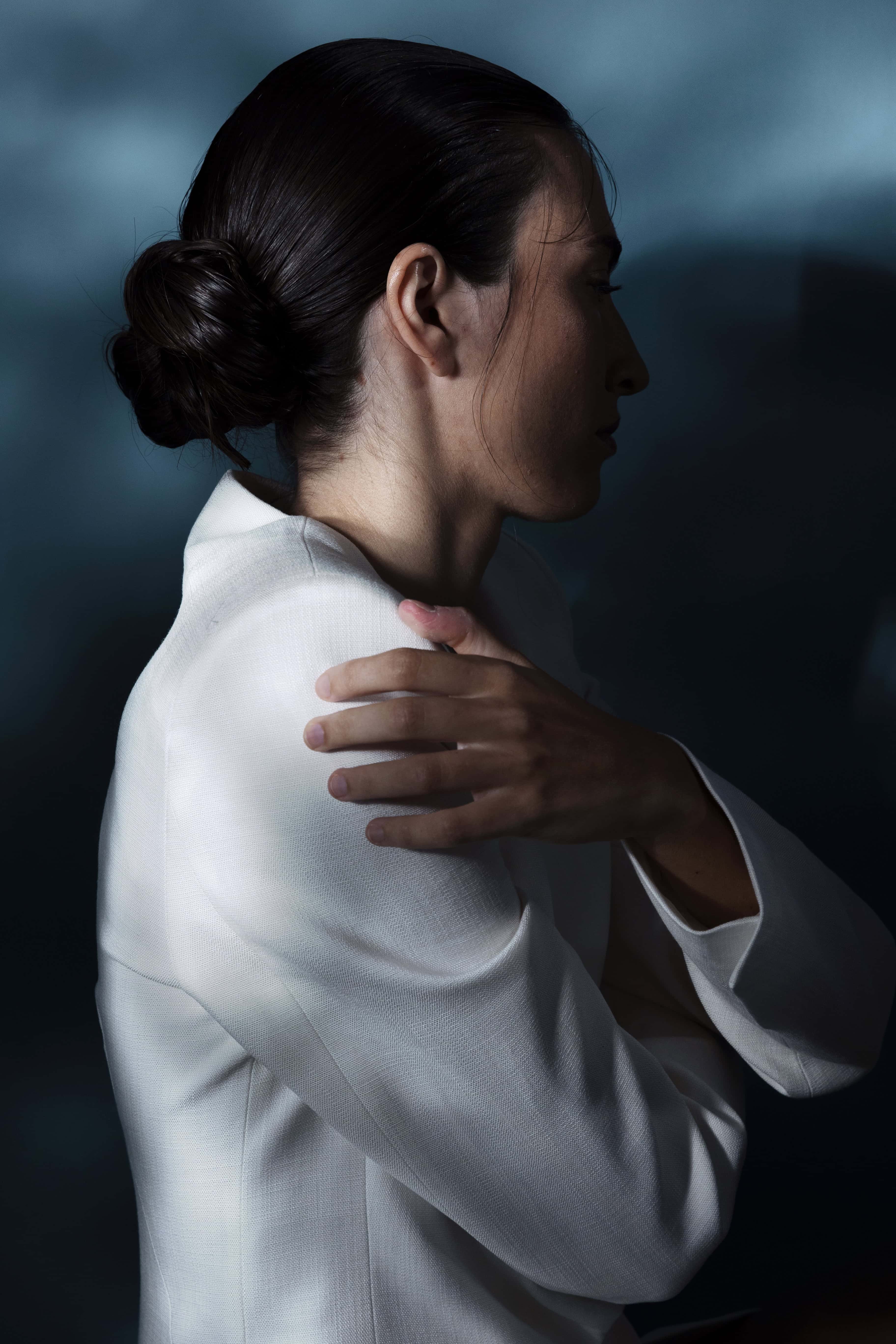 Pohľad zboku na ženu s tmavými vlasmi v drdole a v bielej blúzke pred tmavým pozadím, rukou si pridržiava rameno.