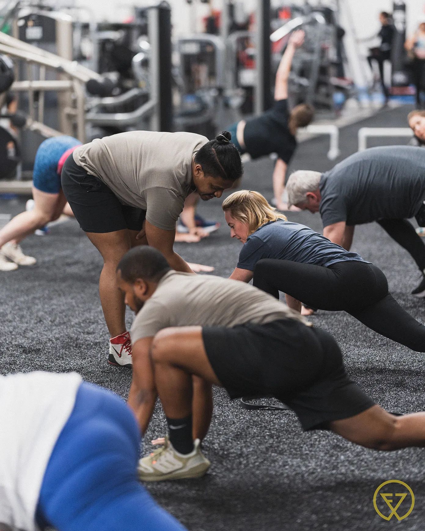Group of people stretching and doing mobility exercises on a gym floor, with an instructor assisting a participant.