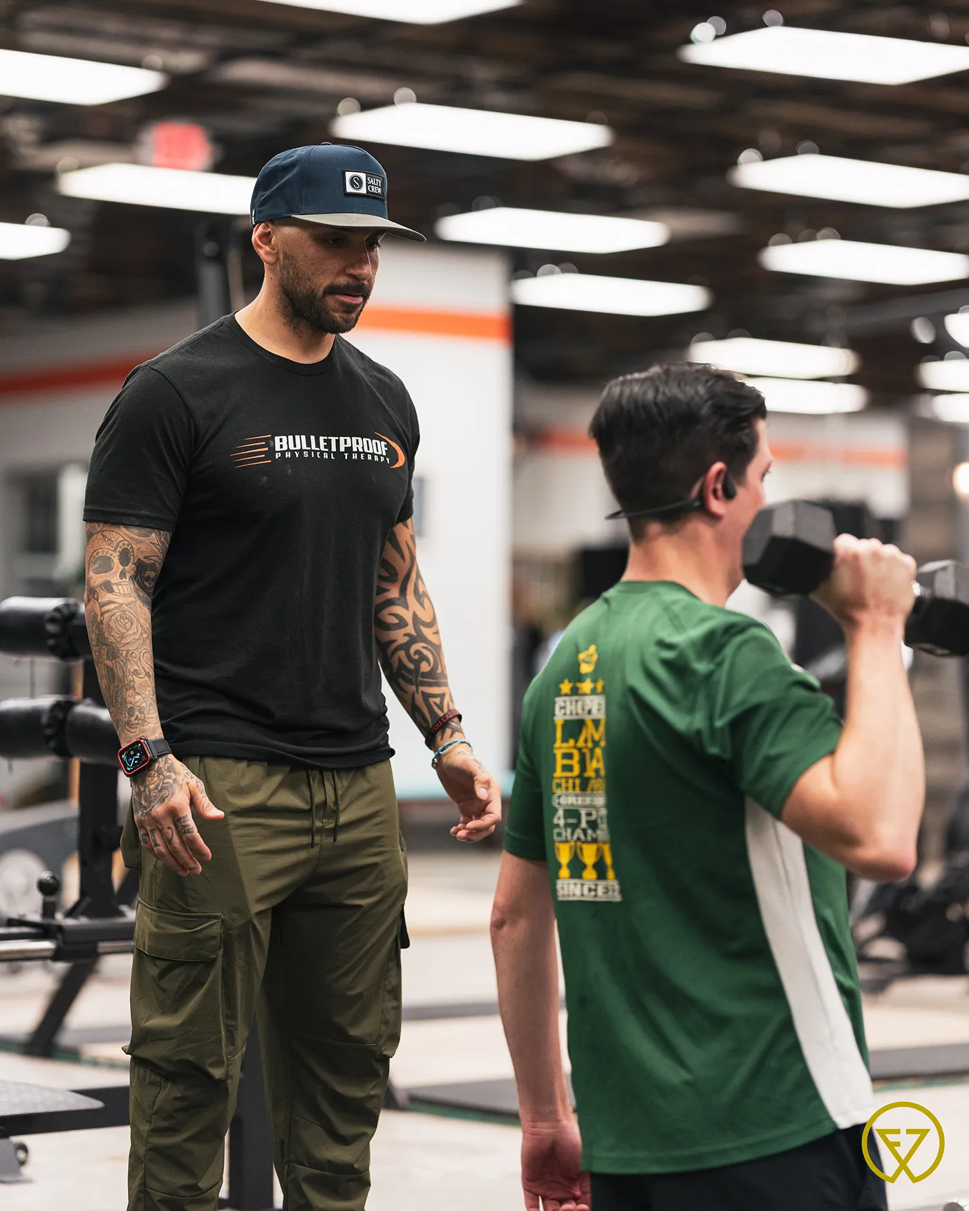 Tattooed male personal trainer in black Bulletproof Physical Therapy shirt coaching a man lifting a dumbbell in a gym.