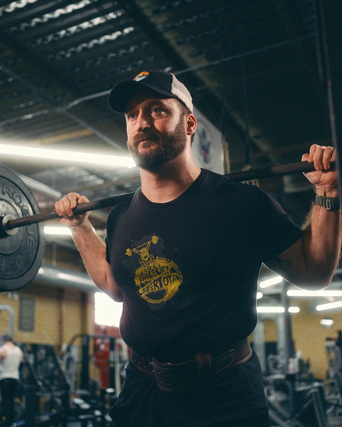 Man with a beard wearing a cap and black t-shirt lifting a barbell on his shoulders in a gym.