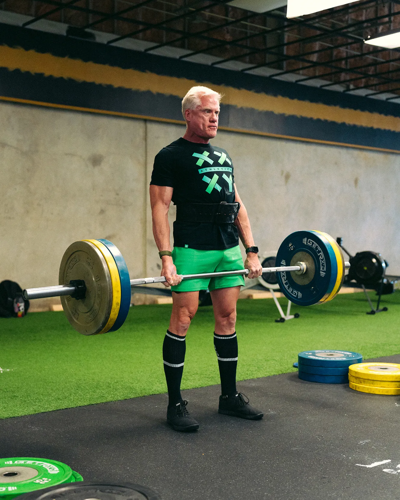 Man lifting a heavy barbell with multiple weight plates in a gym with green turf and weight stacks.