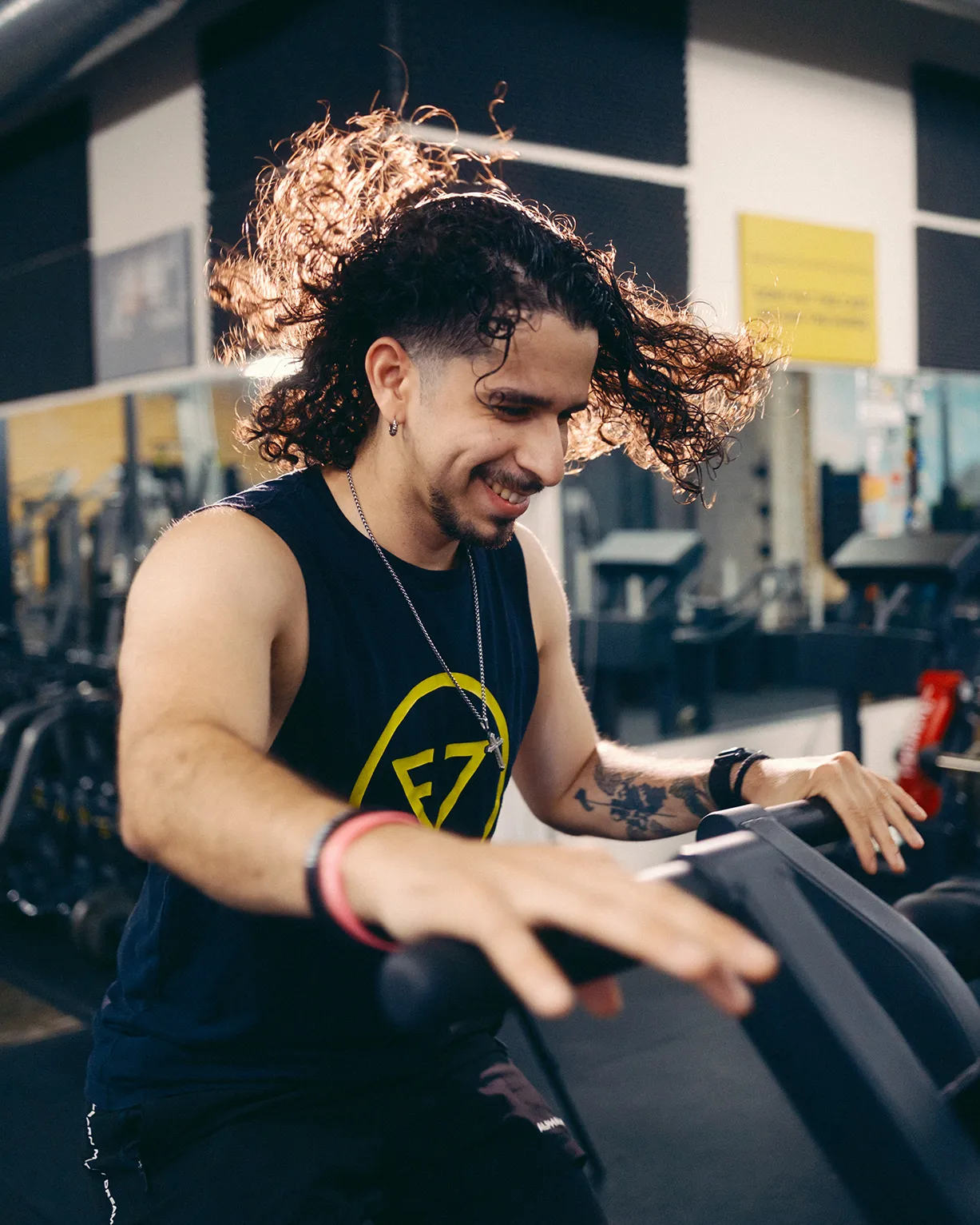 Smiling man with curly hair riding an exercise bike in a gym.