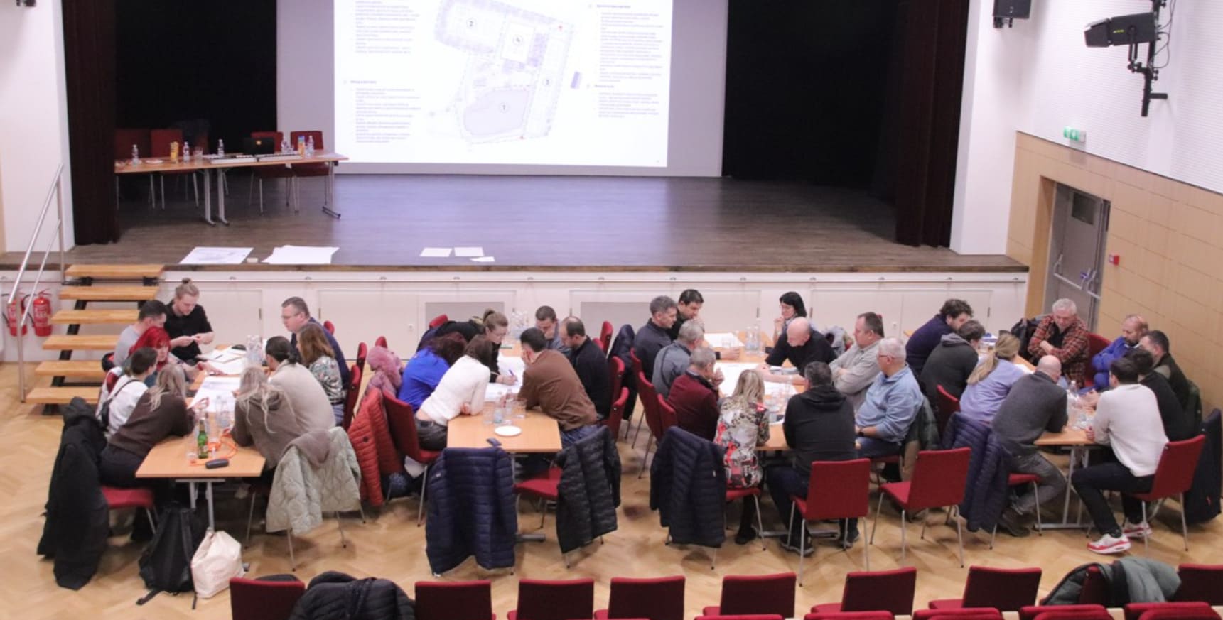 Skupiny lidí sedící u stolů v sále během veřejného setkání nad mapovými podklady, před pódiem s projekční plochou.
