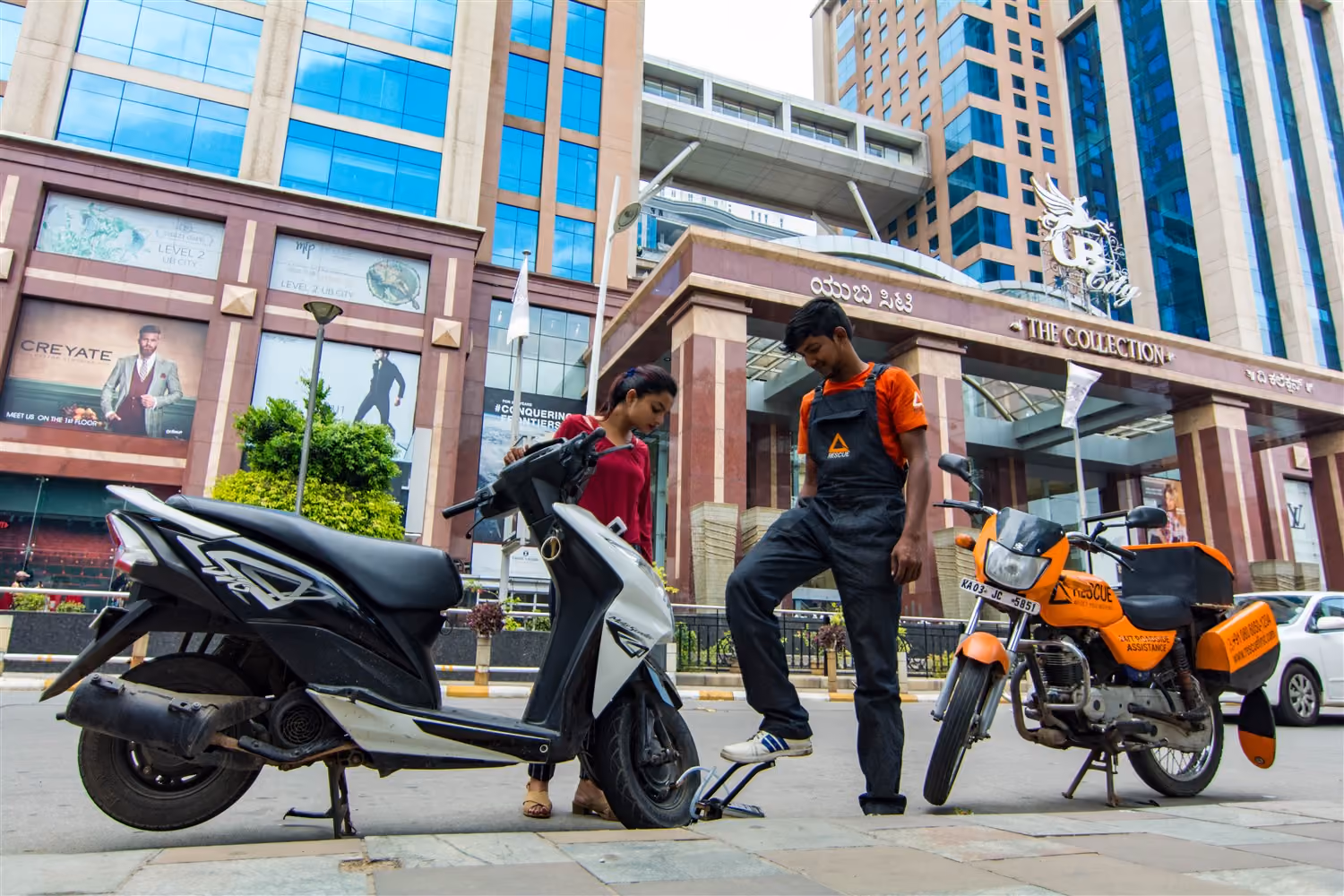 A motorcycle rescue worker repairs a scooter tire on a city sidewalk while a woman looks on.