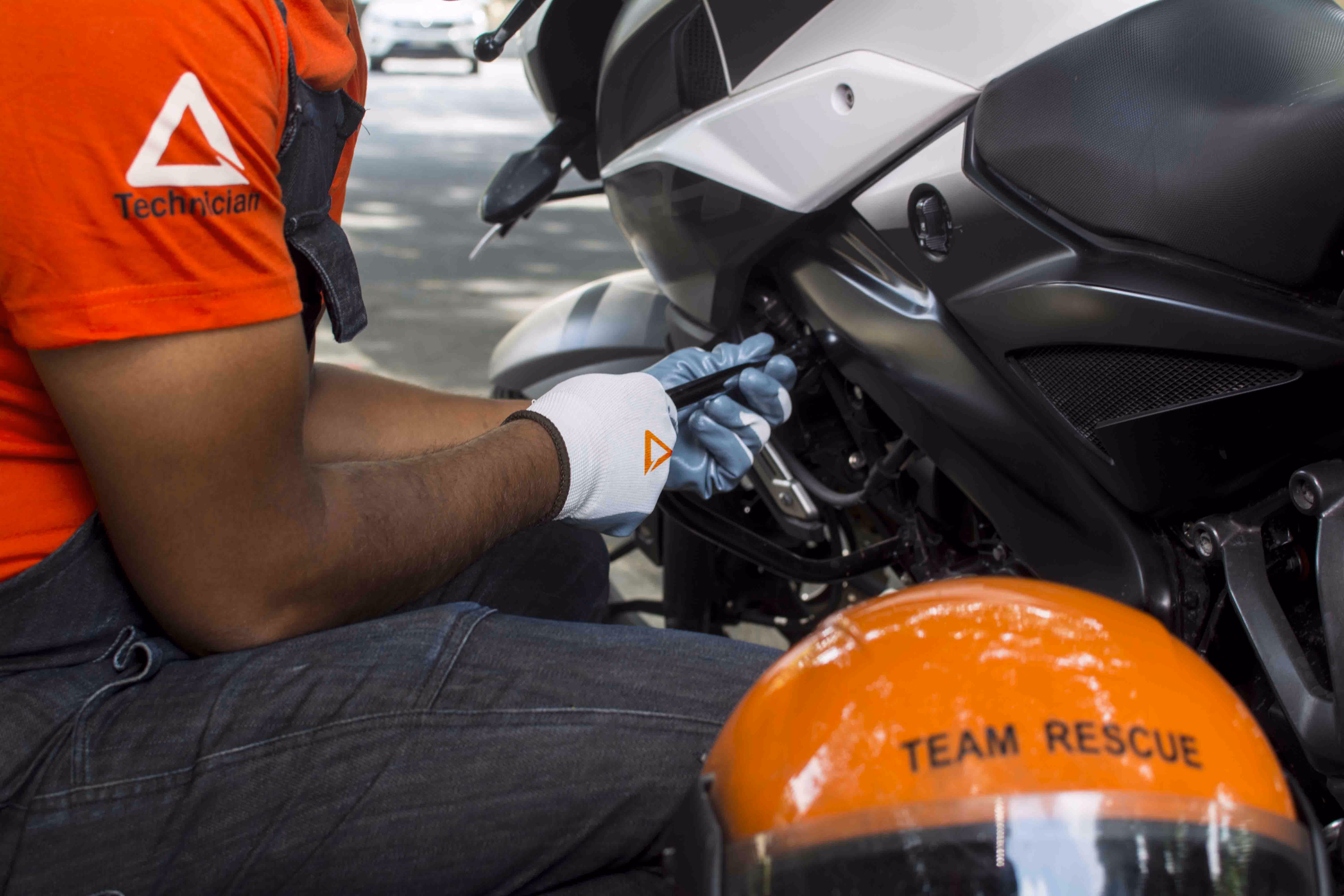 Technician wearing an orange shirt and gloves repairing a black and white motorcycle with an orange helmet labeled 'TEAM RESCUE' in the foreground.