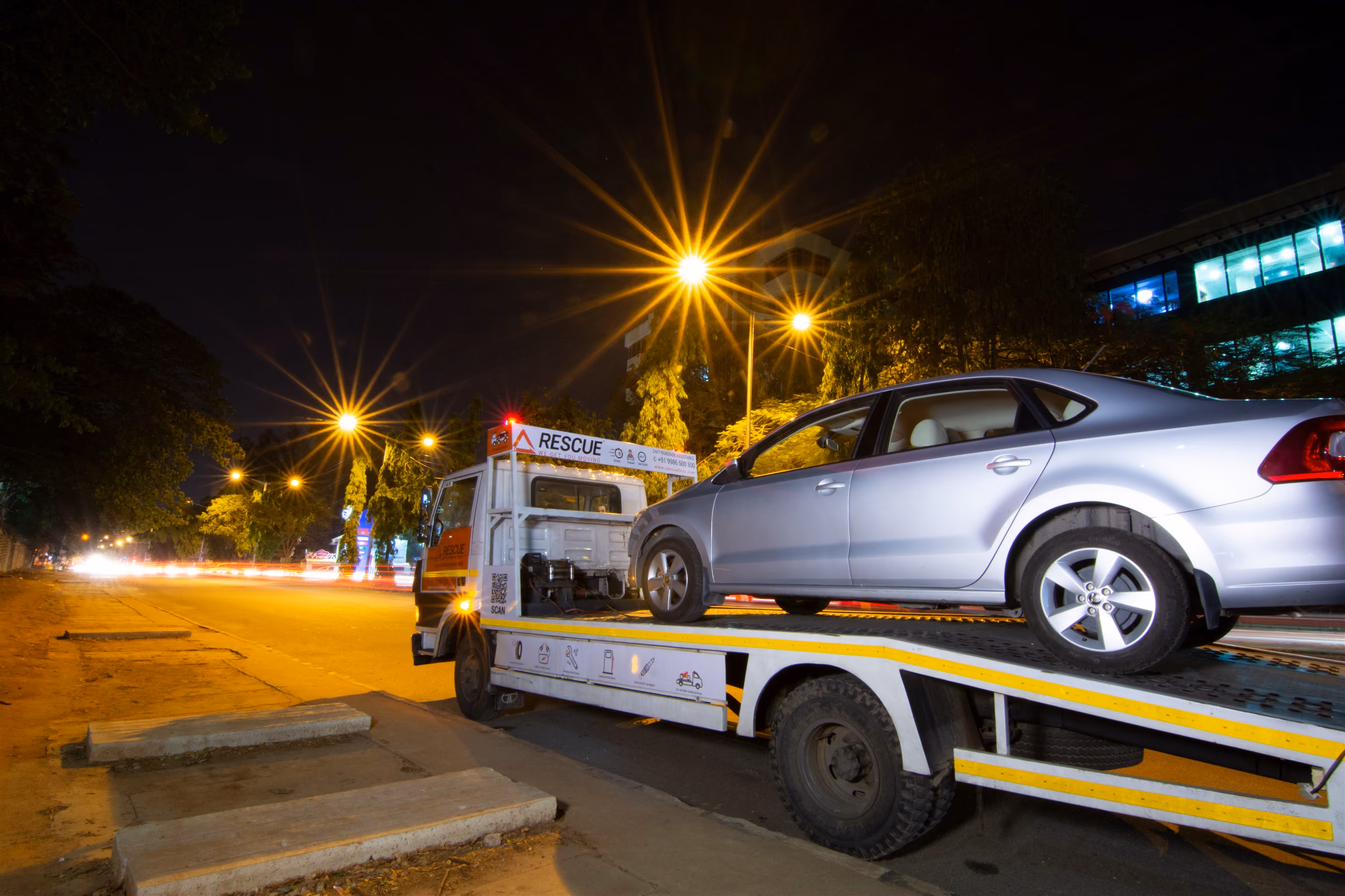 Silver sedan car loaded on a white tow truck at night on a city street with illuminated streetlights.