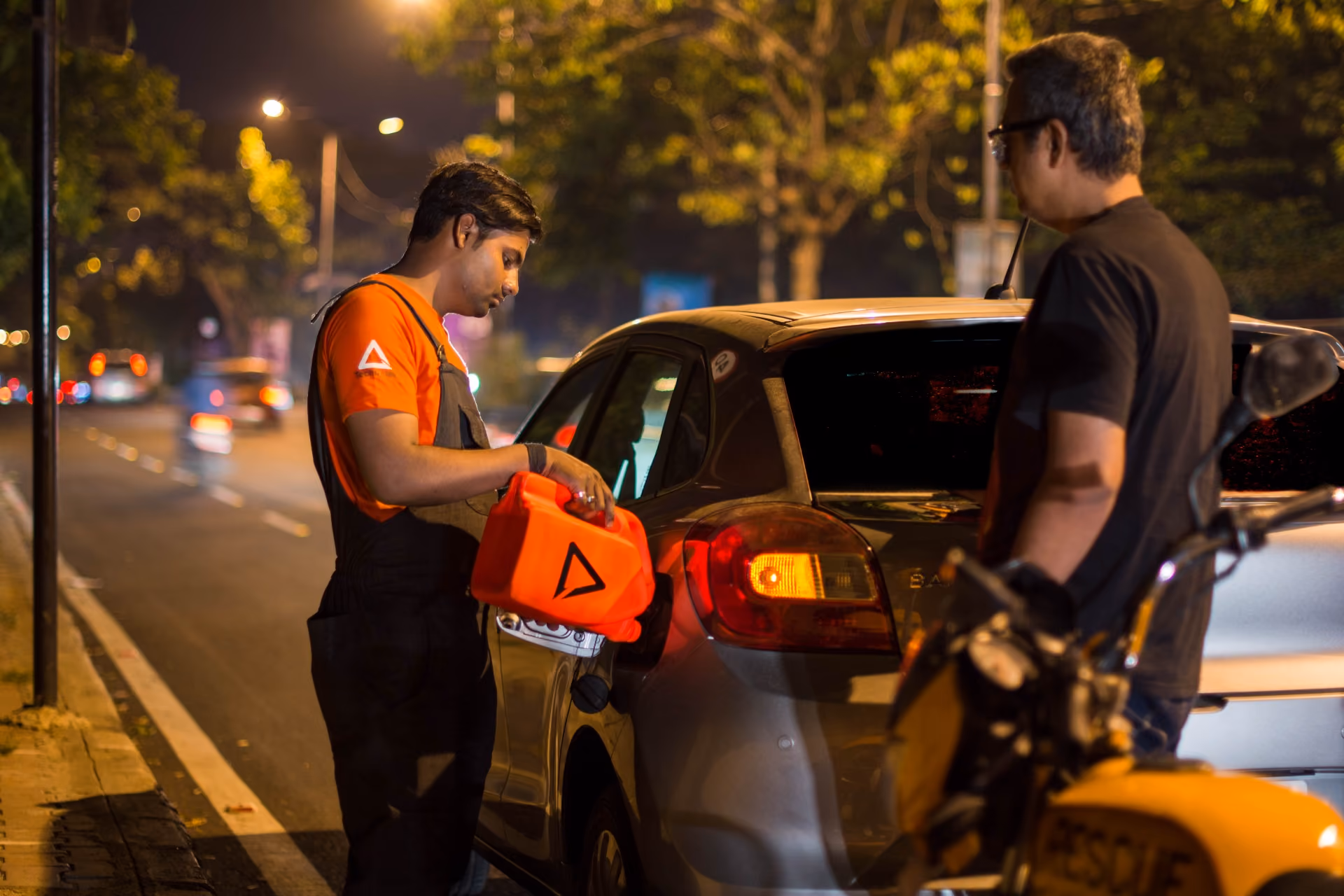 Man in orange shirt refueling a silver car from a red container at night while another man with a motorcycle watches.