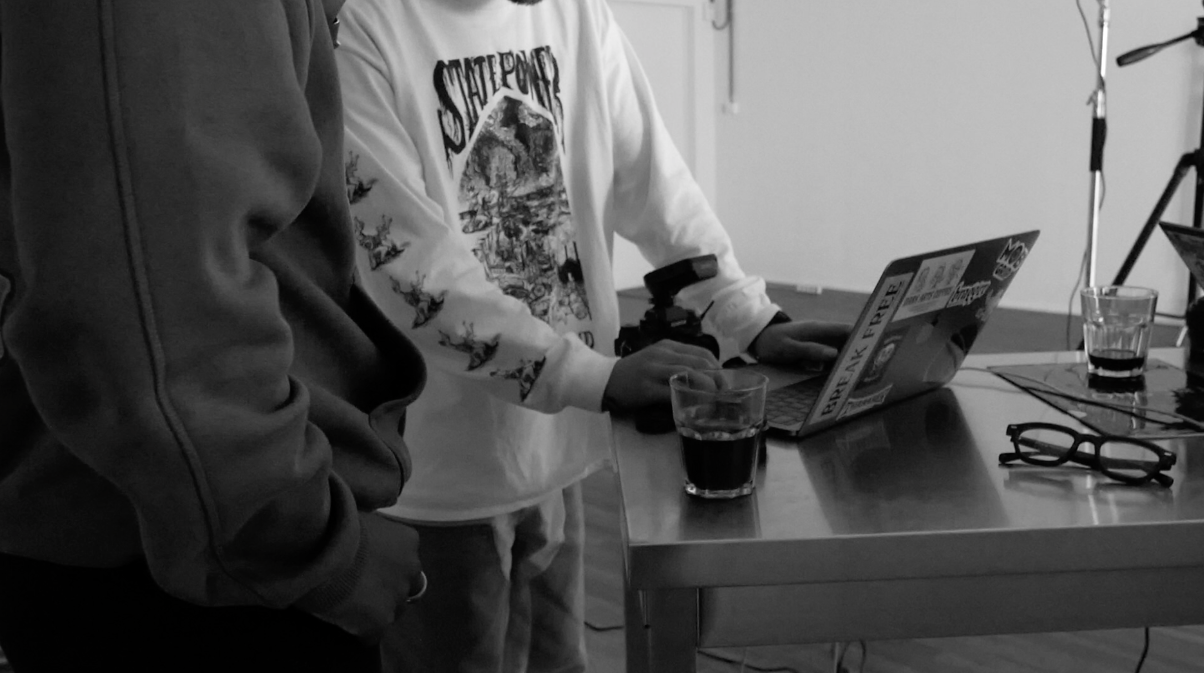 Two people standing by a table with a laptop covered in stickers, glasses, and a camera.