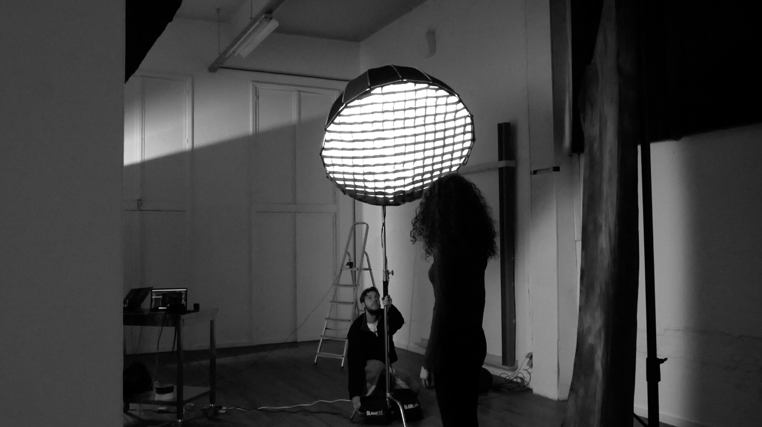 Man adjusting large studio light while woman with curly hair stands facing it in a photo studio.
