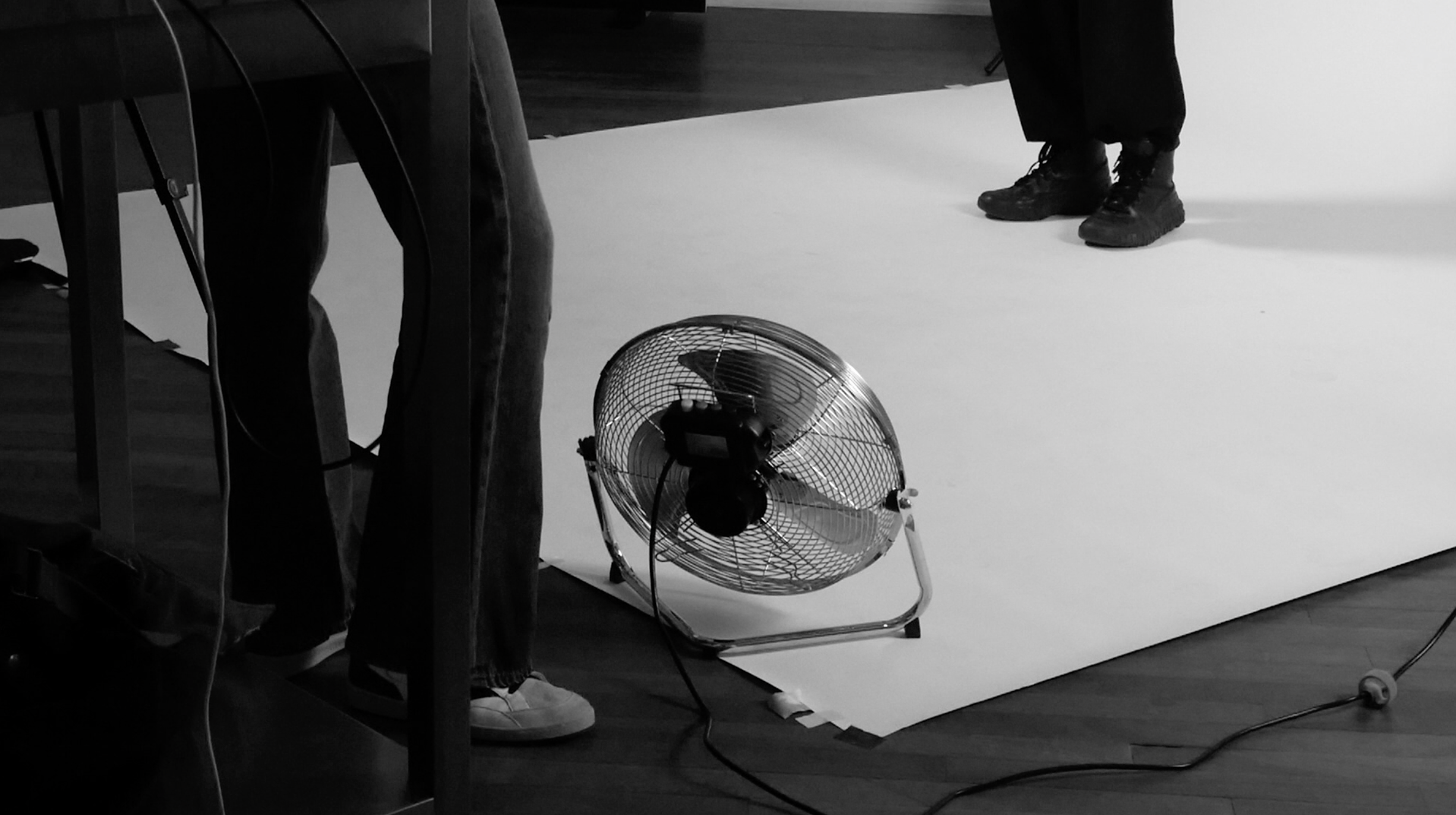 Black and white photo of two people standing near a small floor fan on a wooden floor and white backdrop.