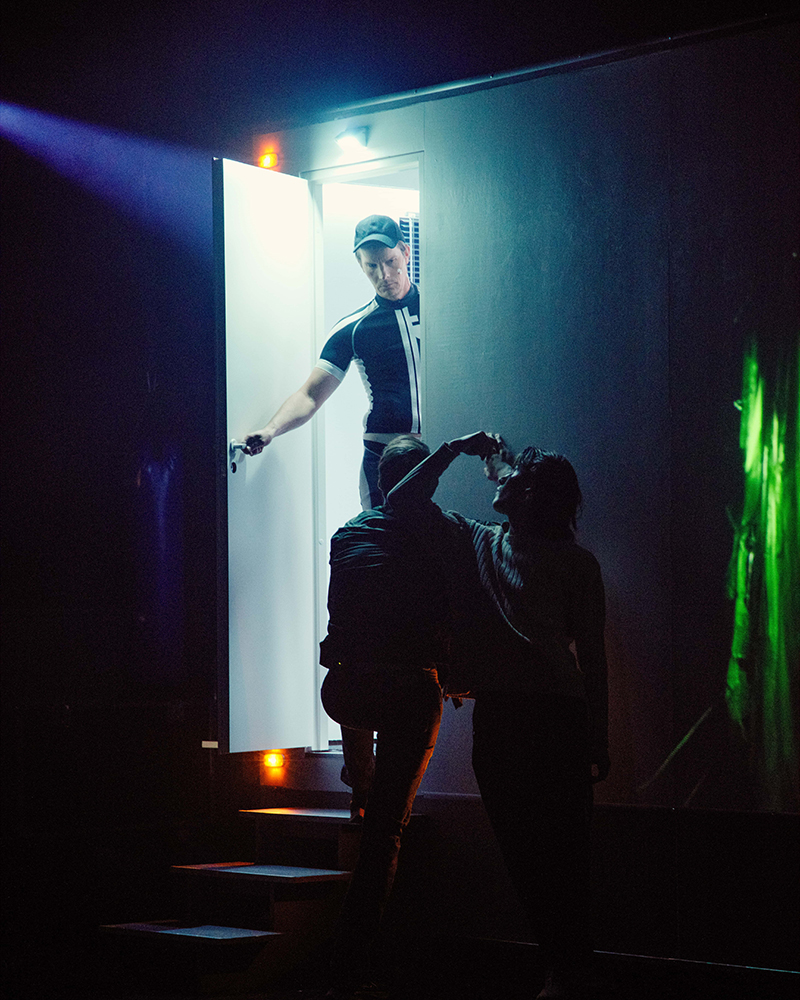 Man in a cap standing in a brightly lit doorway holding the door open for two people drinking and standing in the shadows.