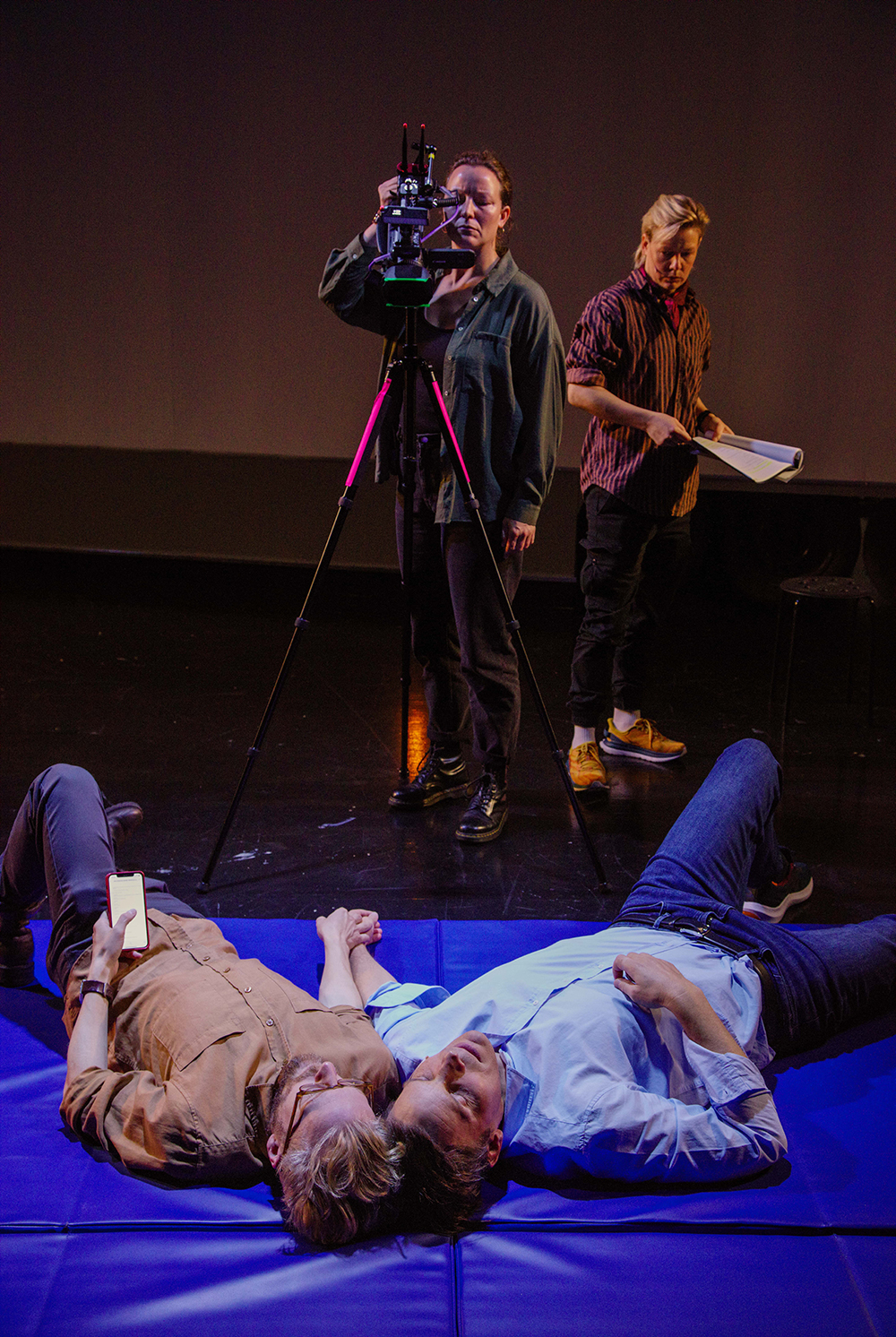 Two people lying on a blue mat holding hands, one looking at a phone, while two others stand behind a camera on a tripod and holding a script.
