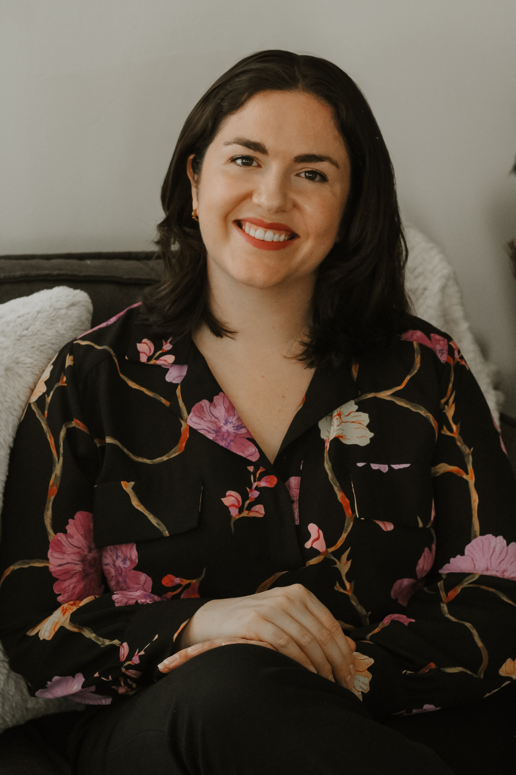 Grace, a smiling woman with long brown hair wearing a black polka dot top, sits in a cozy, warmly lit office with framed credentials and colorful artwork in the background.