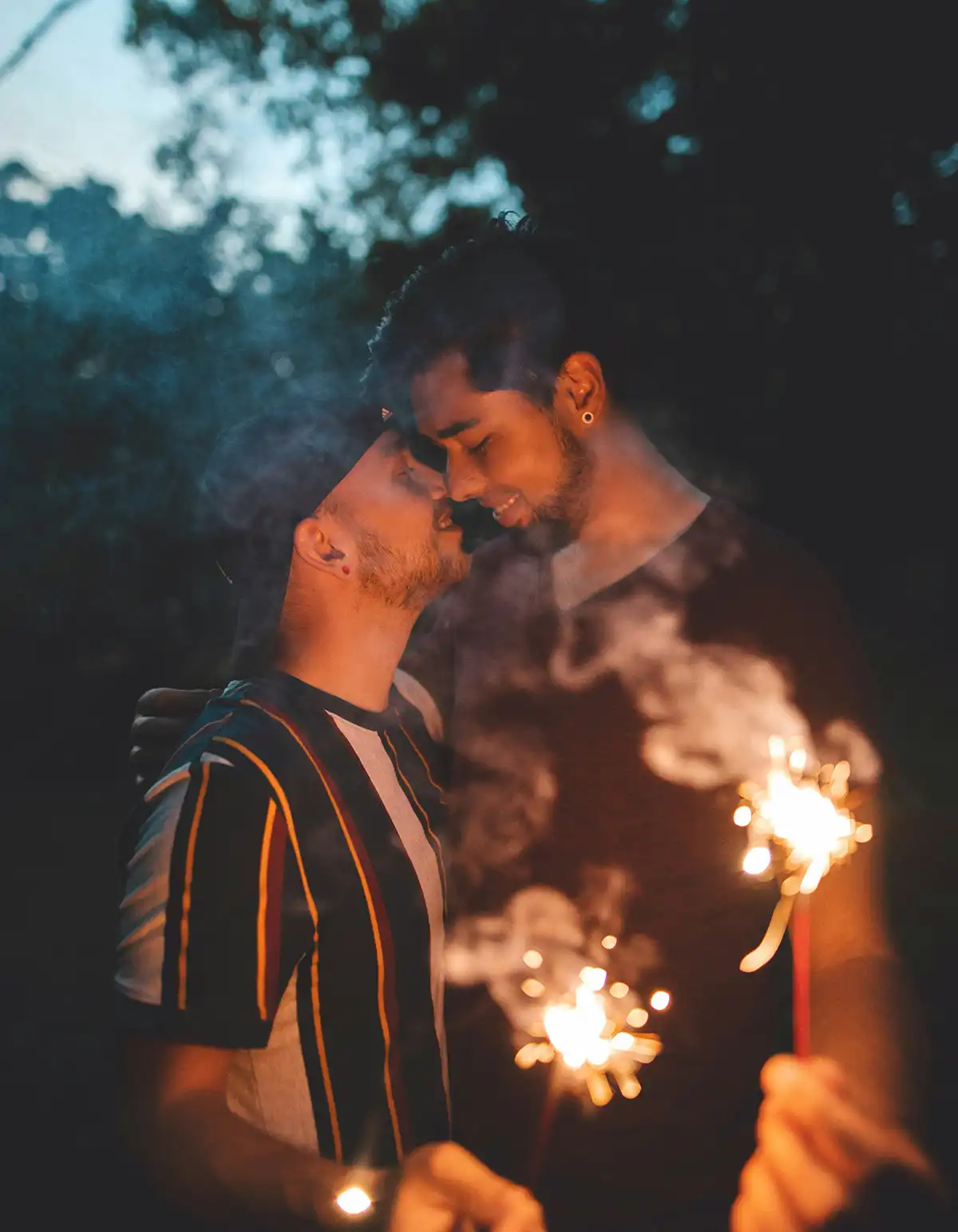 Two men embracing outdoors at dusk, holding sparklers